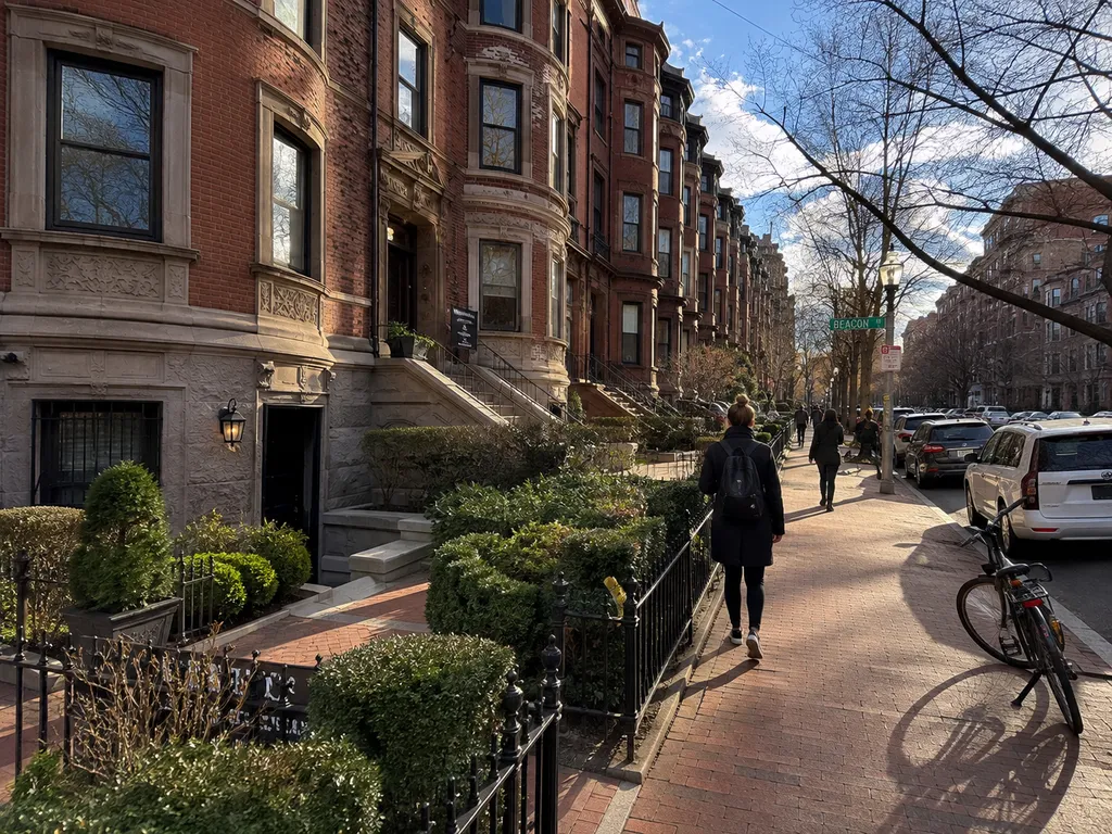 A person walks on a sidewalk with parked cars, buildings, and trees.