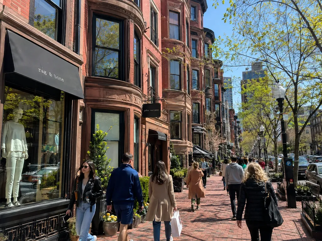 People walking on a brick sidewalk past shops with spring trees.